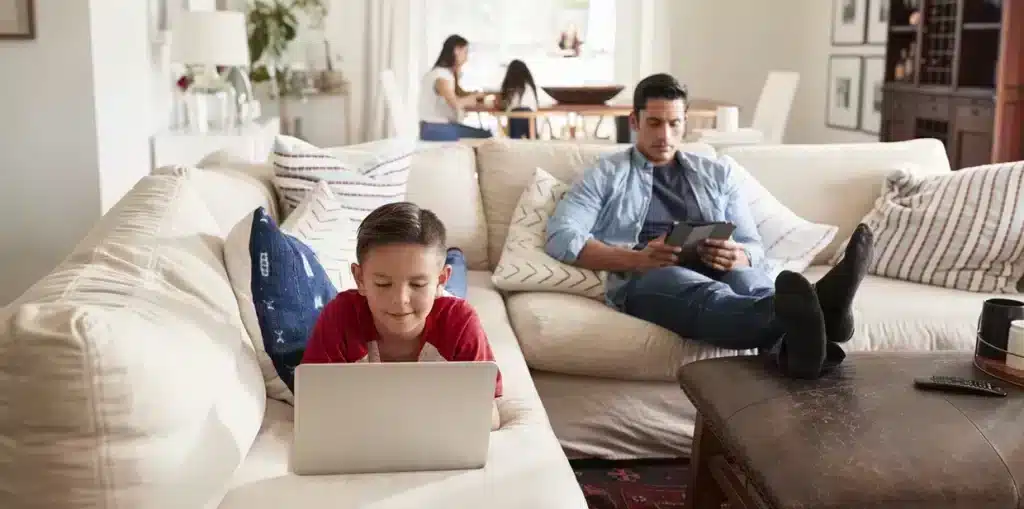 Vue d'un petit garçon allongé sur le canapé avec un ordinateur à côté de son père en train d'utiliser sa tablette